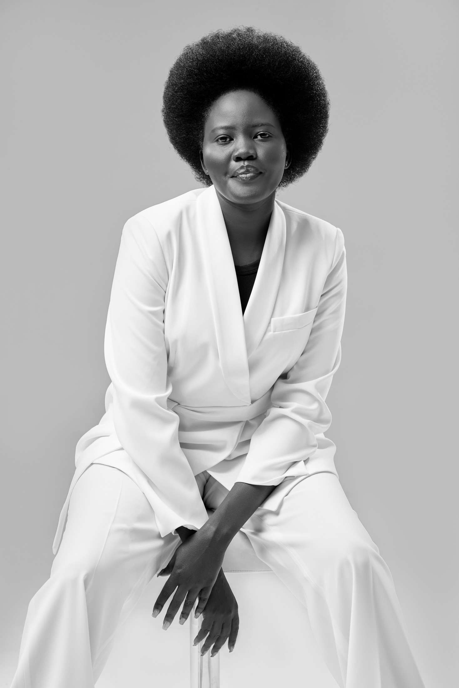Black and white fashion portrait of a woman with an afro in a white suit, seated against a soft grey backdrop