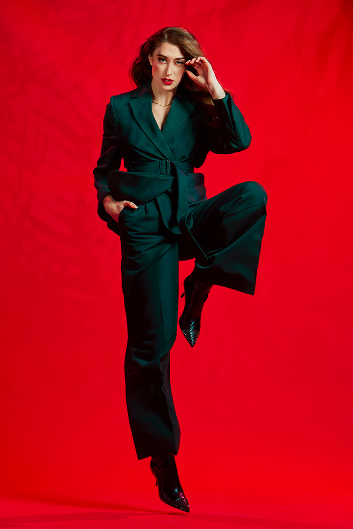 Model in a dark green tailored suit with one leg raised, on a red set