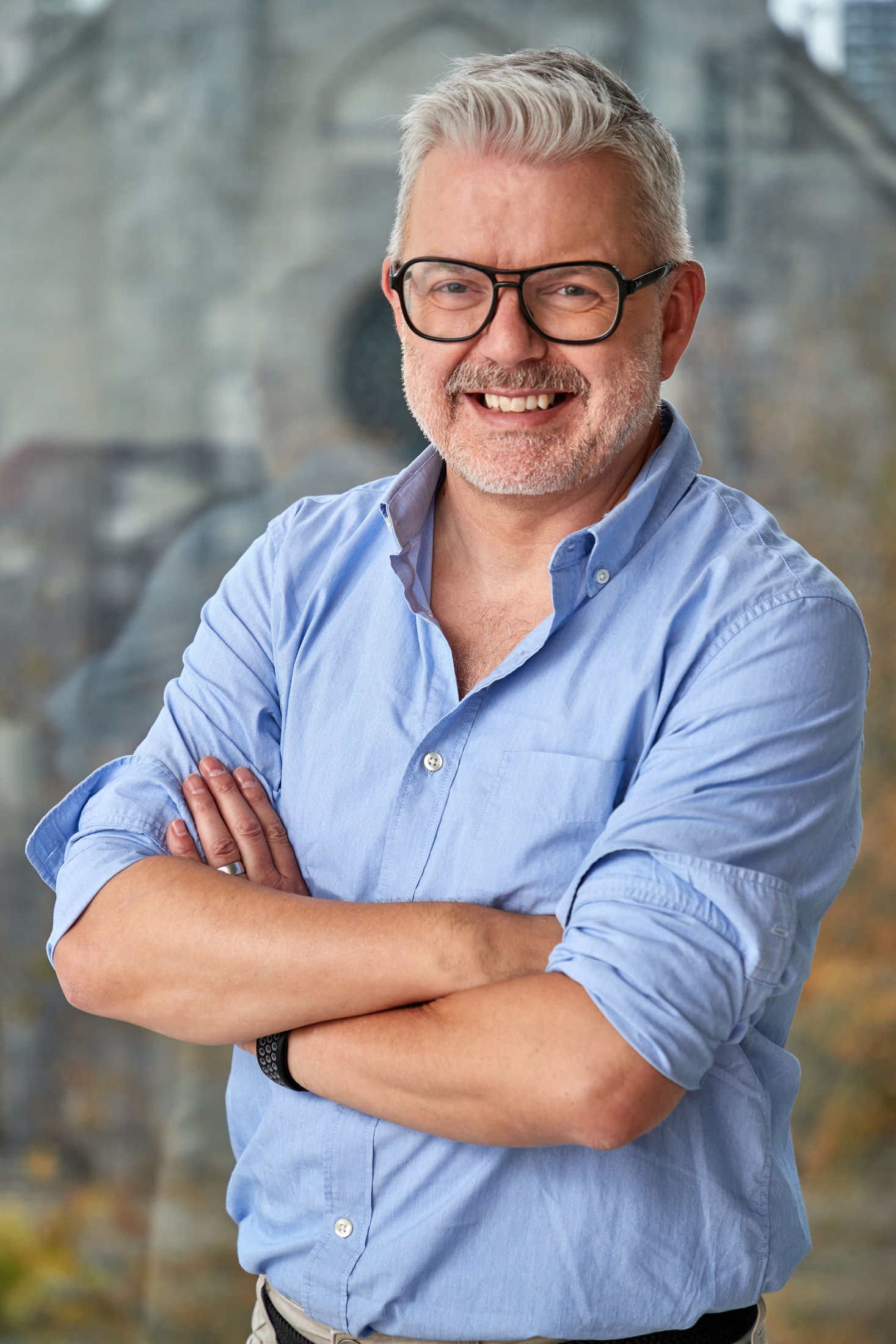 Smiling man with arms crossed in a blue shirt beside a window, personal branding portrait