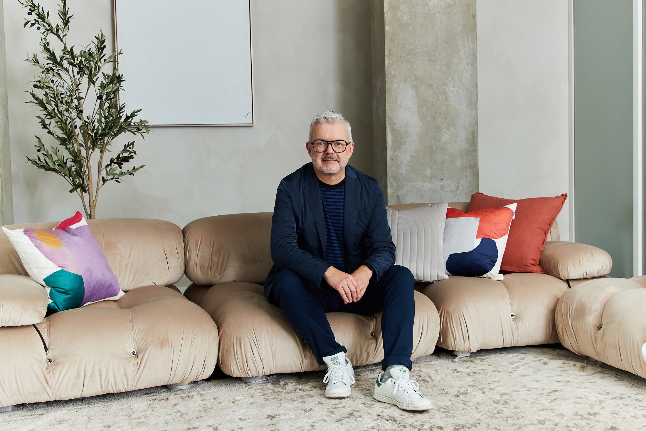 Man in a navy blazer and white sneakers on a large beige sofa, relaxed professional portrait